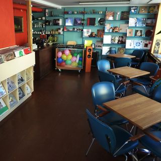 Interior of Cafe Humbug with brown floor, three desks and blue leather chairs on the right, a beige record shelf on the left. In the background a turqouis wall with shelves filled with records and some stash. In front of that ther's a a cabinet with a glass front, filled with colored balloons and turtables on top of it, beside that an orange speaker